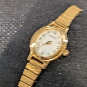 Può includere: Un orologio da polso color oro con quadrante ovale e bianco. L'orologio presenta lancette e indici delle ore dorati. Il cinturino è in metallo flessibile color oro. L'orologio è esposto su una superficie scura e strutturata.