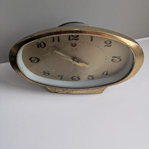 May include: A vintage gold-toned alarm clock with a cream-colored face. The clock has black numerals and hands, and the face is surrounded by a gold-colored bezel. The clock has a rectangular base.