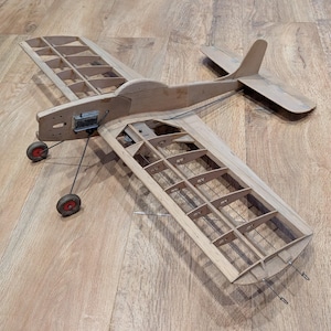 May include: A vintage wooden model aeroplane with a light brown finish. The plane features a detailed wing structure, a tail, and small red and black wheels. The model is likely a collectible or a display piece.