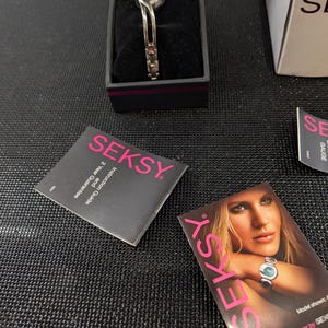Puede incluir: Un reloj Seksy en una caja negra. El reloj tiene una correa plateada con una esfera negra y pequeños detalles rojos. La caja está junto a una tarjeta con la palabra "SEKSY" en rosa.