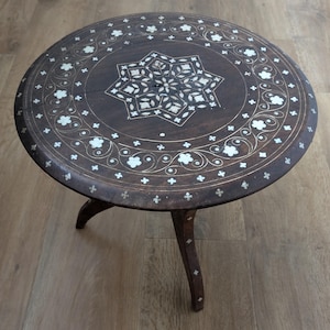 Puede incluir: Mesa auxiliar redonda de madera marrón oscuro con diseño incrustado. La tapa presenta un patrón de estrella central y detalles florales blancos. La mesa tiene tres patas curvas. Aproximadamente 40 cm de diámetro.