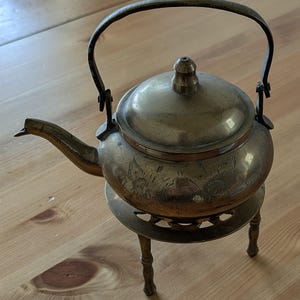 Könnte beinhalten: Eine Messing-Teekanne mit einem verzierten Design steht auf einem Ständer mit drei Beinen. Die Teekanne hat einen Deckel und einen Griff.