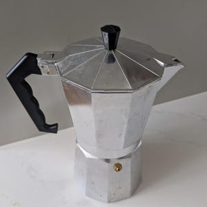May include: A silver-coloured aluminium moka pot with an octagonal base and a faceted upper chamber. It features a black plastic handle, a black knob on the hinged lid, and a small brass-coloured pressure release valve on the side of the lower chamber.