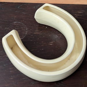 May include: A cream-coloured ceramic vase shaped like a horseshoe with two openings. The vase is designed to hold two separate flower arrangements.