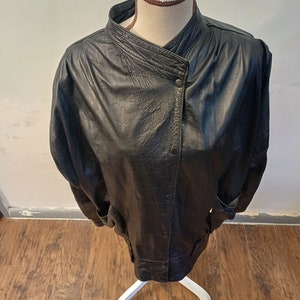 以下が含まれることがあります： スタンドカラーとボタン留めの黒い革のジャケット。ジャケットはゆったりとしたフィット感で、少しオーバーサイズのシルエットです。