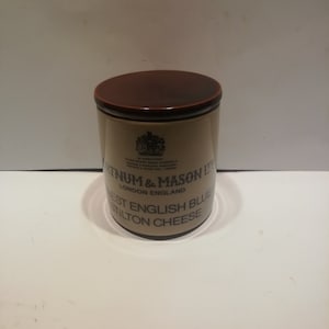 Puede incluir: Un tarro de cerámica marrón y blanco con una tapa marrón. El tarro tiene el texto "Fortnum & Mason Ltd. London England" y "West English Blue Stilton Cheese" impreso en él.