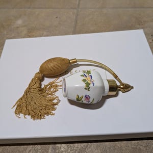 May include: A white porcelain perfume bottle with a floral design and a gold-colored atomizer. The bottle is attached to a gold-colored tube with a tassel.