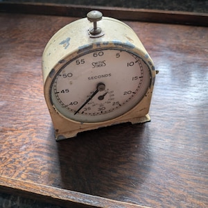 Puede incluir: Un temporizador de cocina vintage blanco con una esfera negra y una perilla plateada. El temporizador está etiquetado como "Smiths Timer" y "Seconds".