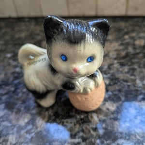 Puede incluir: Una figurita de cerámica negra y blanca de un gato jugando con una pelota. El gato tiene ojos azules y la nariz rosa. La figurita está firmada "Hungría".