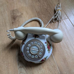 以下が含まれることがあります： クリーム色のヴィンテージロータリー電話。花柄とシルバーのダイヤルが付いています。電話にはコイル状の受話器コードとストレート電源コードが付いています。ダイヤルには数字と「REDIAL/PAUSE」と「THE ROYAL ALBERT TELEPHONE」の文字が表示されています。