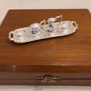 Puede incluir: Un juego de té de plata en una bandeja de plata, dentro de una caja de madera. El juego de té incluye una tetera, una jarra de crema, un azucarero y una taza pequeña.