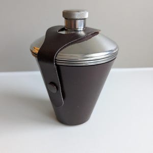 Peut inclure: Une flasque conique en métal avec un sommet argenté poli et une gaine en cuir marron foncé. La gaine comporte une fermeture à bouton-pression et enveloppe la partie supérieure de la flasque, fixée par une sangle passant sur le bouchon à vis.