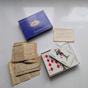 Puede incluir: Un juego de cartas Canasta vintage con una caja azul, una baraja de cartas y recortes de periódicos antiguos. La baraja de cartas incluye una carta de comodín y un seis de diamantes. La caja está etiquetada como "Canasta" y "Waddington".