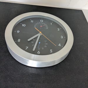 Op de afbeelding: Een ronde weerstationklok met een zilveren frame en een zwarte wijzerplaat. De klok heeft witte cijfers, zilveren wijzers en twee kleinere wijzerplaten. De woorden "WEATHER STATION" staan op de wijzerplaat.