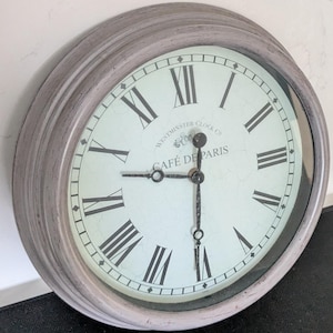 Könnte beinhalten: Eine runde Wanduhr mit einem verwitterten grauen Rahmen und einem cremefarbenen Zifferblatt mit schwarzen römischen Ziffern. In der Mitte steht der Text "WESTMINSTER CLOCK CO" und "CAFÉ DE PARIS" in schwarzer Schrift, mit schwarzen Metallzeigern.