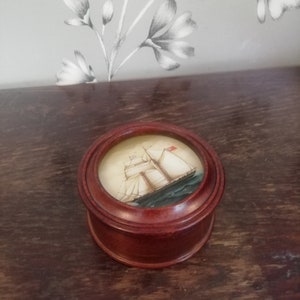 Può includere: Una piccola scatola di legno con un coperchio rotondo. Il coperchio presenta un'immagine dipinta di una nave a vela con vele bianche e una bandiera rosso, bianco e blu.