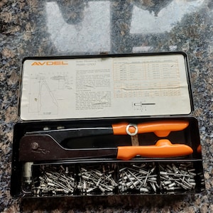 May include: A black and orange Avdel 7598-0100 rivet gun in a metal case with a variety of rivets. The case has a chart with rivet sizes and instructions on how to use the tool.