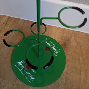 Pode incluir: Porta-copos de metal verde com an&eacute;is de borracha preta. O porta-copos tem uma base redonda com as palavras "Tanqueray" e "Est. London 1830" impressas nele. O porta-copos tem quatro an&eacute;is para segurar copos.