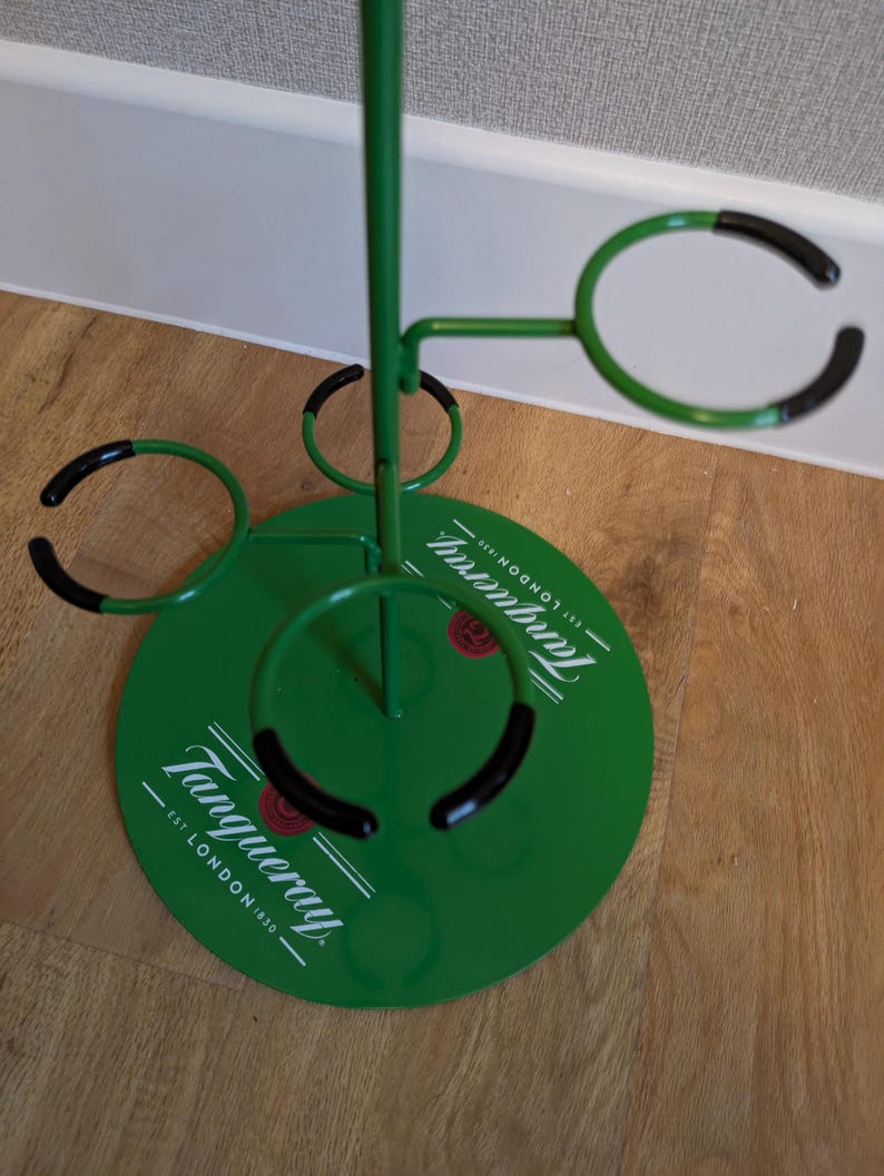 Pode incluir: Porta-copos de metal verde com an&eacute;is de borracha preta. O porta-copos tem uma base redonda com as palavras "Tanqueray" e "Est. London 1830" impressas nele. O porta-copos tem quatro an&eacute;is para segurar copos.