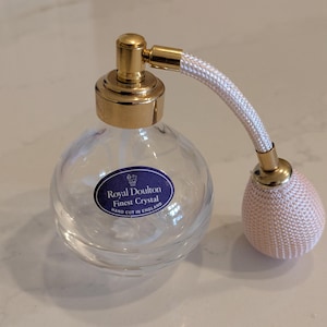 May include: A clear glass atomizer bottle with a gold top and a white fabric pump. The bottle has a blue label that reads "Royal Doulton Finest Crystal Hand Cut in England."
