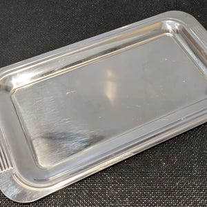 Op de afbeelding: Een rechthoekig zilverkleurig metalen dienblad met een gepolijst, reflecterend oppervlak. Het dienblad heeft afgeronde hoeken en een opstaande rand met een subtiel, lineair geribbeld textuurdetail aan de twee korte zijden.