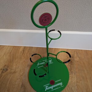 Pode incluir: Porta-garrafas de metal verde com base redonda e quatro an&eacute;is para segurar garrafas. A base tem o logotipo da Tanqueray e o texto "EST. LONDON 1830".