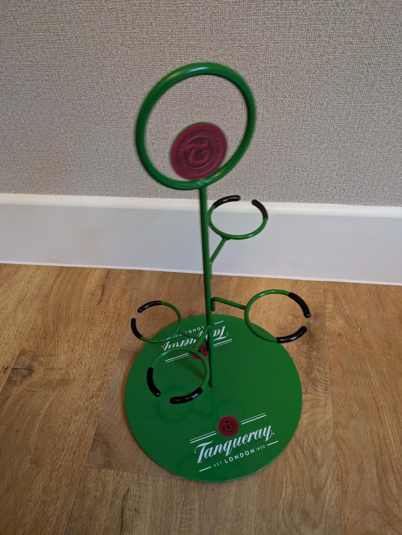 Pode incluir: Porta-garrafas de metal verde com base redonda e quatro an&eacute;is para segurar garrafas. A base tem o logotipo da Tanqueray e o texto "EST. LONDON 1830".