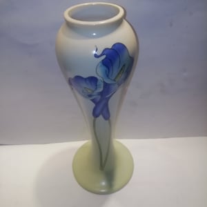 May include: A white ceramic vase with a tall, slender neck and a flared base. The vase is decorated with two blue and white flowers.