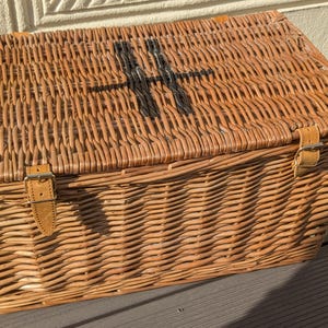 May include: Wicker chest with a rectangular shape and a hinged lid. The chest is woven from light brown wicker and features a black cross painted on the lid. Leather straps with buckles secure the lid.