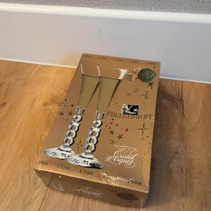 Puede incluir: Una caja dorada con dos copas de champán con tallos de eslabones de cadena plateados. La caja tiene las palabras "Millennium" y "Cristal d'Arques" impresas. La caja está sobre una superficie de madera.