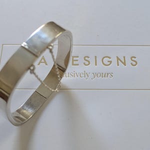 以下が含まれることがあります： ヒンジ留め具とチェーンのディテールが付いたシルバーのバングルブレスレット。ブレスレットは、「A Designs Exclusively Yours」というテキストが印刷された白い表面の上に置かれています。