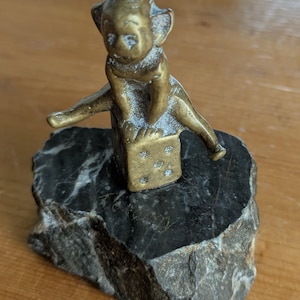Può includere: Statuetta vintage in ottone di un elfo seduto sopra un dado, montata su una base di pietra scura e strutturata. L'elfo ha un cappello a punta e appoggia le braccia sul dado. Il dado mostra i numeri 1, 2 e 3. La base in pietra è larga circa 10 cm.
