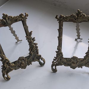 May include: Two antique bronze-colored picture frames with ornate, scrolling details. The frames have a rectangular shape with decorative embellishments and small feet for standing. The frames are empty, ready to display photos or artwork.
