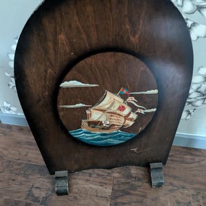 May include: A wooden fireplace screen featuring a painted image of a ship with sails and flags. The screen has a dark brown finish and three legs.