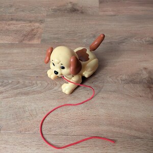 Op de afbeelding: Een gele en bruine speelgoedhond met een rood touwtje vastgemaakt aan zijn bek. De hond staat op een houten vloer.