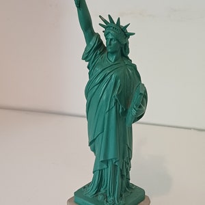 May include: A teal-colored Statue of Liberty figurine with a gold-colored torch. The statue stands on a light gray base, with the words "MADE IN USA" visible. The figurine is a miniature replica of the iconic landmark.