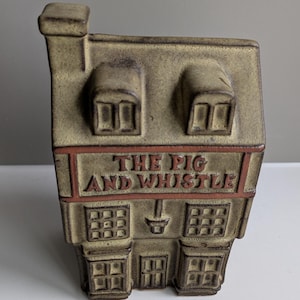 May include: A ceramic figurine shaped like a pub, with the text "THE PIG AND WHISTLE" in red lettering. The building is tan with brown accents, featuring windows, doors, and a chimney. The overall design resembles a traditional English pub.