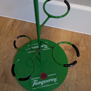 Pode incluir: Porta-garrafas de metal verde com an&eacute;is de borracha preta para segurar garrafas. A base do porta-garrafas &eacute; verde e tem o logotipo da Tanqueray com o texto "EST. LONDON 1830".