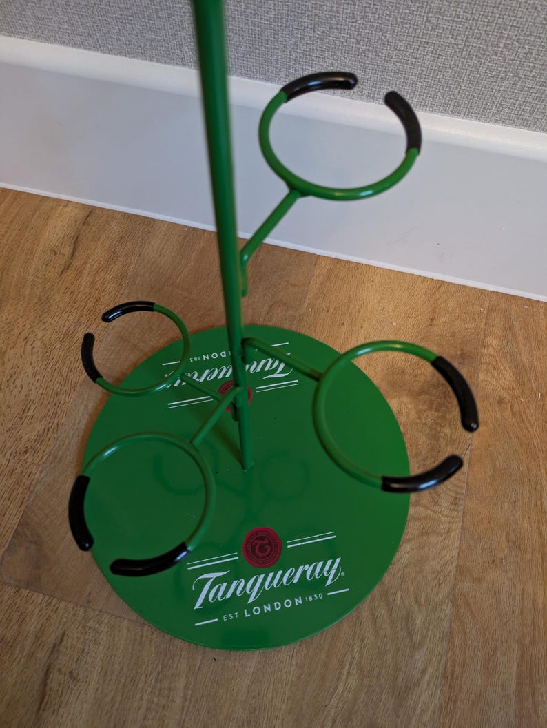 Pode incluir: Porta-garrafas de metal verde com an&eacute;is de borracha preta para segurar garrafas. A base do porta-garrafas &eacute; verde e tem o logotipo da Tanqueray com o texto "EST. LONDON 1830".