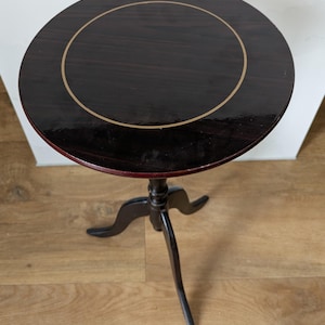 May include: A small, round, dark brown side table with a glossy finish. The tabletop features a gold-coloured ring around the edge. The table has a three-legged base, and the legs are curved. The table is approximately 61 cm tall.