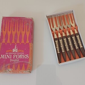Op de afbeelding: Een doos met zes mini-vorkjes van gebronsd metaal met houten handvatten. De doos is roze en oranje en heeft de tekst "Six Bronze Plated Mini Forks Disneyland". De vorkjes zitten in een witte doos met het woord "Disneyland" op elk handvat.