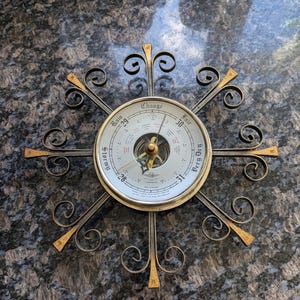 May include: A vintage brass barometer with a black metal frame and a decorative design. The barometer has a round face with a white background and black markings. The face is labeled with the words "Change", "Stormy", "Fair", "Very Dry", and "Rain".