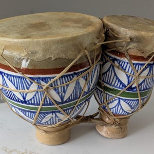 May include: A set of two connected bongo drums featuring a hand-painted ceramic body with blue, green, and brown geometric patterns. The drum heads are made of natural tan animal hide, secured with a decorative network of thin, light-colored twine.