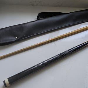May include: A two-piece pool cue featuring a light brown wooden shaft and a black butt section with a textured diamond-patterned grip and a white ferrule, accompanied by a black faux leather carrying case with a handle and a metal snap closure.