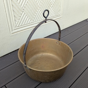 May include: A round brass pot with a smooth interior and a weathered exterior surface. The pot features a dark, arched metal handle attached to the sides with rivets, and a small circular loop at the top center of the handle for hanging.