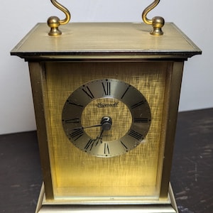 Puede incluir: Un reloj rectangular dorado con una esfera negra y números romanos. El reloj tiene una manija en la parte superior y dice "Ingersoll" en la esfera.