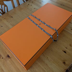 May include: An orange rectangular box with a black trim and a patterned ribbon tied around it. The box is on a wooden surface. The ribbon has a repeating pattern with the brand name.