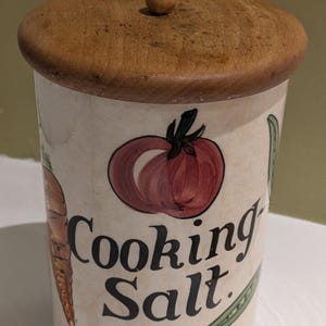 Peut inclure: Un pot à sel de cuisine en céramique avec un couvercle en bois. Le récipient est décoré d'illustrations de légumes, dont une tomate, une carotte et des haricots verts. Les mots "Cooking Salt" sont imprimés sur le devant.