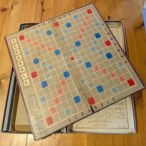 Pode incluir: Um tabuleiro de jogo Scrabble vintage com um esquema de cores castanho e bege. O tabuleiro é feito de cartão e tem uma grelha de 15x15 quadrados. Os quadrados são marcados com cores e letras diferentes, indicando diferentes valores de pontos para cada letra. O tabuleiro é dobrado ao meio e tem a palavra "SCRABBLE" impressa na frente e atrás.