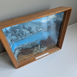 Op de afbeelding: Een ingelijst landschapsdiorama met een rivierlandschap met bergen en bomen. Het kunstwerk is ingekaderd in een houten frame. De tekst onderaan luidt "Ben Lettery Mountain, Connemara."